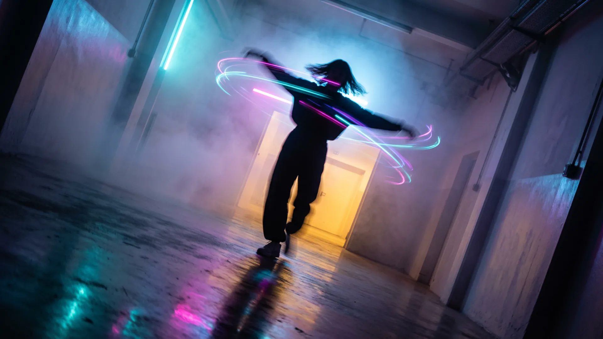 In a burst of uninhibited movement, a figure is captured mid-spin in a corridor of light and mist. The photography freezes the action at its peak, the person's arms flung wide, their head tilted back in a moment of pure, kinetic release. This rapid twirl has visibly disturbed the dense, colored fog, creating eddies and vortices of cyan, magenta, and violet light around their silhouetted form. The composition is vibrant and chaotic, a stark contrast to a static scene. Light from a warm, yellow-orange portal ahead illuminates the swirling mist from one direction, while a cool blue glow from behind adds depth and complexity to the color palette. The floor, a dark mirror, reflects a blurred, chaotic version of the scene—a painterly smear of motion and color. The camera angle is slightly tilted, a Dutch angle that enhances the feeling of disequilibrium and exhilaration, capturing a fleeting, joyful rebellion against the quiet, contemplative nature of the space
